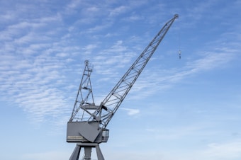 A large metal crane with a long arm extends into a partly cloudy blue sky. The crane has a series of structural supports and a hook hanging from a cable at the end of the arm. The base is stationary and appears sturdy.