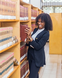 A person in a navy suit is smiling while pulling a large book from a wooden bookshelf filled with similar volumes. The setting appears to be a library with rows of bookshelves and a tiled floor.