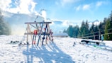A snowy landscape with a rack holding multiple pairs of skis and ski poles. Nearby, a snowboard is resting in the snow. Trees line the background as mountains and a partly cloudy sky stretch expansively, creating a serene winter scene. A skier dressed in winter attire is seen in the distance.