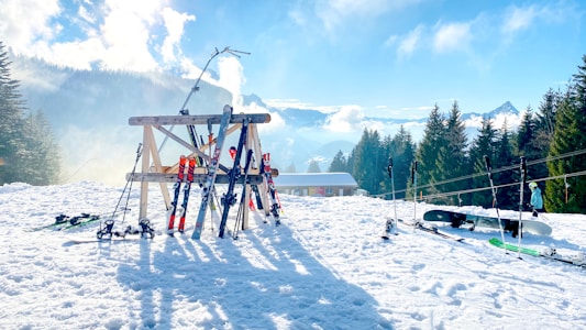 A snowy landscape with a rack holding multiple pairs of skis and ski poles. Nearby, a snowboard is resting in the snow. Trees line the background as mountains and a partly cloudy sky stretch expansively, creating a serene winter scene. A skier dressed in winter attire is seen in the distance.