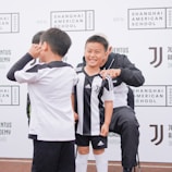 A group of children wearing black and white soccer uniforms are standing on a sports field. One child is smiling broadly, while another makes a playful gesture. They are near a backdrop with the Shanghai American School and Juventus Academy logos.