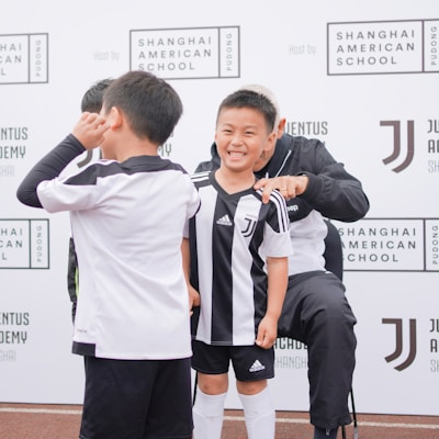 A group of children wearing black and white soccer uniforms are standing on a sports field. One child is smiling broadly, while another makes a playful gesture. They are near a backdrop with the Shanghai American School and Juventus Academy logos.