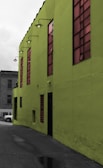 A vibrant green building with large tinted windows stands prominently. The building's exterior features industrial-style lamps mounted along the upper edge. A small portion of a gray concrete alleyway is visible, along with a parked car in the background.