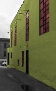 A vibrant green building with large tinted windows stands prominently. The building's exterior features industrial-style lamps mounted along the upper edge. A small portion of a gray concrete alleyway is visible, along with a parked car in the background.