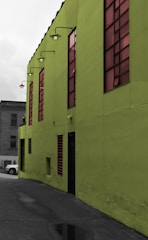 A vibrant green building with large tinted windows stands prominently. The building's exterior features industrial-style lamps mounted along the upper edge. A small portion of a gray concrete alleyway is visible, along with a parked car in the background.