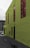 A vibrant green building with large tinted windows stands prominently. The building's exterior features industrial-style lamps mounted along the upper edge. A small portion of a gray concrete alleyway is visible, along with a parked car in the background.