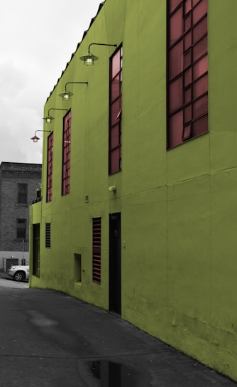 A vibrant green building with large tinted windows stands prominently. The building's exterior features industrial-style lamps mounted along the upper edge. A small portion of a gray concrete alleyway is visible, along with a parked car in the background.
