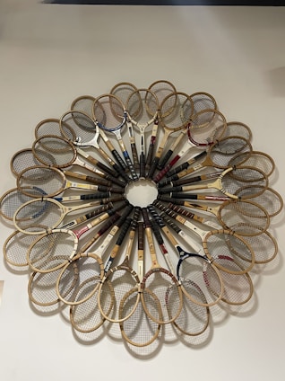 A circular arrangement of vintage wooden tennis rackets forms a symmetrical pattern against a light-colored wall. Each racket is positioned with the handle pointing outward, showcasing the unique designs and strings of the rackets. The overall composition resembles a floral or radial design.