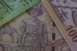 Banknotes overlapping each other, featuring an intricate design with historical figures, buildings, and decorative patterns. The notes display a range of vignettes, including a figure in military attire and architectural elements.