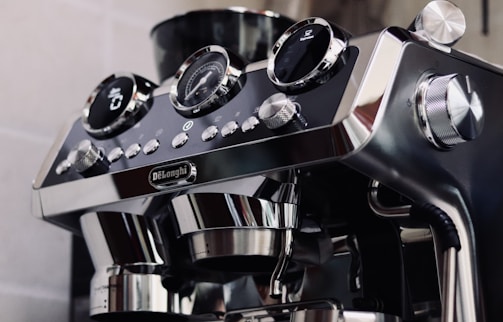 A sleek and modern espresso machine with a shiny, metallic finish. It features multiple knobs and buttons, including a digital display and analog gauges, likely for precision in brewing coffee. The brand name is prominently displayed on the machine.