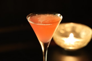 A vibrant pink cocktail is elegantly served in a martini glass, set against a dark background. The drink has a frothy surface with visible reflections on the glass. To the right, a candle flickers inside a glass holder, adding a warm, ambient glow to the scene.