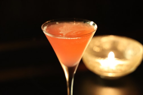 A vibrant pink cocktail is elegantly served in a martini glass, set against a dark background. The drink has a frothy surface with visible reflections on the glass. To the right, a candle flickers inside a glass holder, adding a warm, ambient glow to the scene.