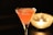 A vibrant pink cocktail is elegantly served in a martini glass, set against a dark background. The drink has a frothy surface with visible reflections on the glass. To the right, a candle flickers inside a glass holder, adding a warm, ambient glow to the scene.