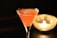A vibrant pink cocktail is elegantly served in a martini glass, set against a dark background. The drink has a frothy surface with visible reflections on the glass. To the right, a candle flickers inside a glass holder, adding a warm, ambient glow to the scene.