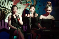 Three women are seated side by side, dressed in black attire. They appear to be performing music, as they each have a microphone. The middle woman is smiling, while the other two have neutral expressions. One of the women is playing a Yamaha keyboard. The background features a vibrant, colorful mural with musical notes, instrument designs, and partial text 'ONE'.