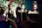 Three women are seated side by side, dressed in black attire. They appear to be performing music, as they each have a microphone. The middle woman is smiling, while the other two have neutral expressions. One of the women is playing a Yamaha keyboard. The background features a vibrant, colorful mural with musical notes, instrument designs, and partial text 'ONE'.