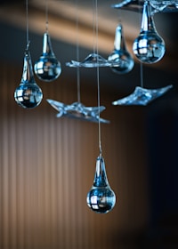 Several glass ornaments, including teardrop-shaped and abstract forms, are hanging from thin wires against a blurred background. The glass is highly polished, reflecting light and the surroundings.