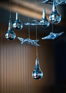 Several glass ornaments, including teardrop-shaped and abstract forms, are hanging from thin wires against a blurred background. The glass is highly polished, reflecting light and the surroundings.