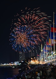Vibrant fireworks fill the night sky with bursts of red and blue hues, illuminating a large crowd below. People are gathered along a waterfront, with numerous colorful flags lined up on poles. Buildings and a well-lit structure are visible in the background, creating a festive atmosphere.