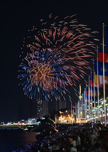 Vibrant fireworks fill the night sky with bursts of red and blue hues, illuminating a large crowd below. People are gathered along a waterfront, with numerous colorful flags lined up on poles. Buildings and a well-lit structure are visible in the background, creating a festive atmosphere.