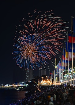 Vibrant fireworks fill the night sky with bursts of red and blue hues, illuminating a large crowd below. People are gathered along a waterfront, with numerous colorful flags lined up on poles. Buildings and a well-lit structure are visible in the background, creating a festive atmosphere.