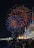 Vibrant fireworks fill the night sky with bursts of red and blue hues, illuminating a large crowd below. People are gathered along a waterfront, with numerous colorful flags lined up on poles. Buildings and a well-lit structure are visible in the background, creating a festive atmosphere.