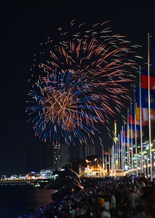Vibrant fireworks fill the night sky with bursts of red and blue hues, illuminating a large crowd below. People are gathered along a waterfront, with numerous colorful flags lined up on poles. Buildings and a well-lit structure are visible in the background, creating a festive atmosphere.