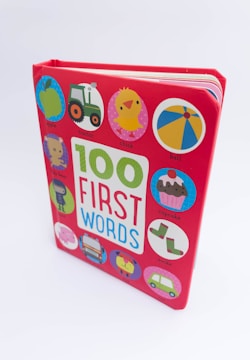 A children's board book with a bright red cover featuring the title '100 First Words' in large colorful letters. Surrounding the title are various circular illustrations including an apple, tractor, chick, ball, cupcake, socks, bus, and car, each labeled with their corresponding word.