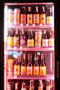A refrigerator filled with an assortment of bottled and canned beers with colorful labels, neatly arranged on shelves. The labels feature a variety of colors and graphics, including images and text.
