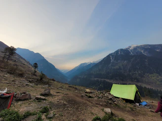 A serene mountain campsite at dawn with a glowing tent pitched near pine trees and a snow-capped peak in the background.