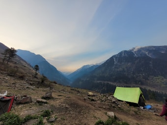 A serene mountain landscape with a wide valley surrounded by snow-capped peaks. A green tent is pitched on rocky ground near a few scattered trees, suggesting a camping site in the wilderness. The sky has a soft gradient from blue to pale yellow, indicating either dawn or dusk.