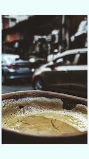 A close-up view of a frothy beverage in a cup, possibly chai, with strands of saffron floating on the surface. The background is blurred, showing a busy street with parked cars and urban structures.