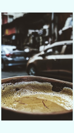 A close-up view of a frothy beverage in a cup, possibly chai, with strands of saffron floating on the surface. The background is blurred, showing a busy street with parked cars and urban structures.