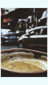 A close-up view of a frothy beverage in a cup, possibly chai, with strands of saffron floating on the surface. The background is blurred, showing a busy street with parked cars and urban structures.