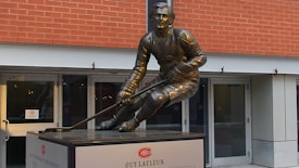 A bronze statue of a hockey player in action is displayed in front of a red brick building. The player is crouched down, holding a hockey stick, and wearing traditional hockey attire. The pedestal of the statue features signage including the name Guy Lafleur.