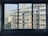 A view of apartment buildings through a four-paned window. The buildings feature multiple stories with balconies and air conditioning units. Laundry hangs from some of the balconies, and the buildings are a beige color. In the background, there is a taller building and some trees visible at the base of the apartment structures.