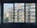 A view of apartment buildings through a four-paned window. The buildings feature multiple stories with balconies and air conditioning units. Laundry hangs from some of the balconies, and the buildings are a beige color. In the background, there is a taller building and some trees visible at the base of the apartment structures.