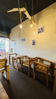 A cozy interior featuring wooden tables and chairs with a rustic design. The walls display framed photographs, and there is a unique chandelier made of driftwood and exposed bulbs hanging from the ceiling. Natural light enters through a glass door, providing a warm and inviting atmosphere.