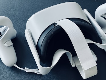 A virtual reality headset is placed on a textured dark surface. The headset is flanked by two handheld controllers, all in a sleek white design. The straps and buttons are visible, suggesting functionality and interactivity.