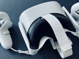 A virtual reality headset is placed on a textured dark surface. The headset is flanked by two handheld controllers, all in a sleek white design. The straps and buttons are visible, suggesting functionality and interactivity.