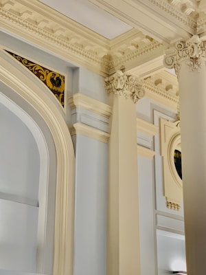 Intricately designed architectural details featuring ornate columns and decorative molding. The ceiling displays elaborate cornices and the walls have subtle curved archways. A hint of stained glass detail is visible in the top right corner.