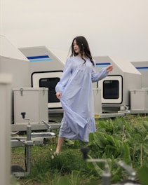 A woman in a long, light blue dress stands gracefully among greenery. In the background, there are several modern, light gray trailers with blue accents.