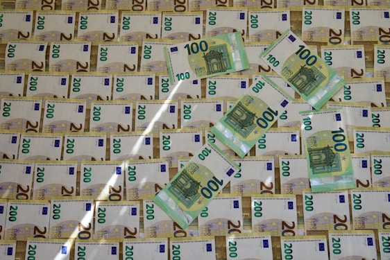 A collection of Euro banknotes, predominantly featuring 100 and 200 euro notes, arranged in a slightly overlapping pattern on a surface. The positioning of the notes creates a sense of abundance and wealth.