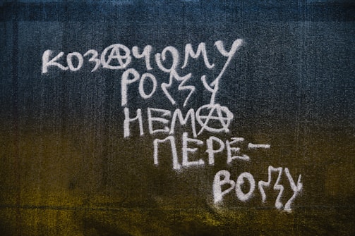 White graffiti text with anarchist symbols is painted on a dark textured wall. The text is written in a stylized font with elements of irregularity and creativity, incorporating the anarchy sign within the letters.