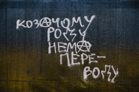 White graffiti text with anarchist symbols is painted on a dark textured wall. The text is written in a stylized font with elements of irregularity and creativity, incorporating the anarchy sign within the letters.