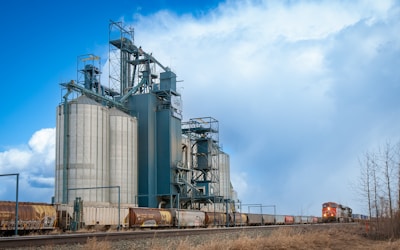 A large industrial grain elevator structure stands beside a railway track. The sky is partly cloudy with patches of blue, creating a contrast with the towering silos of the facility. A train with several freight cars is visible, moving past the silos. Sparse, leafless trees appear in the foreground, indicating a late autumn or early spring setting.