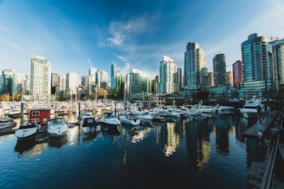 A city skyline with numerous modern high-rise buildings under a clear blue sky. In the foreground, a marina filled with various boats, including yachts and small vessels, is visible. The water in the marina reflects the buildings and the vibrant sky.