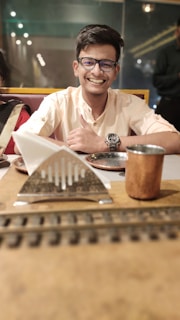 A person with short dark hair and glasses is smiling warmly from behind a table. They are wearing a light-colored shirt with rolled-up sleeves and a watch on the wrist. In front of them are various dining items, including a tissue holder, a metallic cup, and a blurred foreground element that resembles a miniature railway track. The background is softly blurred, suggesting an indoor setting with ambient lighting.