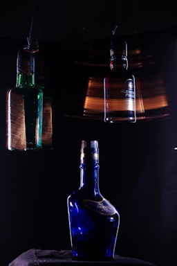 In a dimly lit setting, three glass bottles with different designs are prominently featured against a dark background. The central object is a blue bottle with a reflective surface, sitting on a dark fabric. Above it, two other bottles, one green and one more neutral in color, are suspended, each slightly blurred, suggesting motion or a long exposure effect. The lighting creates dramatic highlights and reflections on the bottles, enhancing their colors.