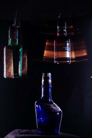 In a dimly lit setting, three glass bottles with different designs are prominently featured against a dark background. The central object is a blue bottle with a reflective surface, sitting on a dark fabric. Above it, two other bottles, one green and one more neutral in color, are suspended, each slightly blurred, suggesting motion or a long exposure effect. The lighting creates dramatic highlights and reflections on the bottles, enhancing their colors.